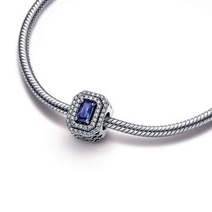 May include: A silver charm bracelet with a blue and white charm featuring a square-shaped blue gemstone surrounded by clear cubic zirconia stones.