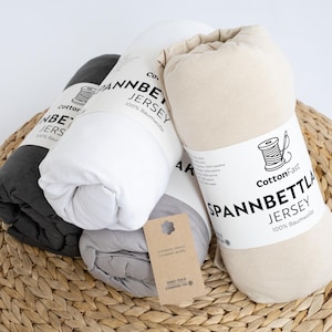 May include: Four rolled jersey knit bed sheets in shades of grey, white, and beige. Each sheet is labelled with the text "CottonFast SPANNBETTLA JERSEY 100% Baumwolle". The sheets are displayed on a woven wicker surface.