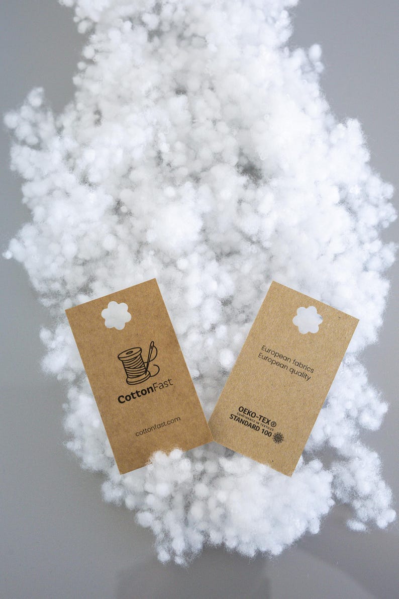 May include: A close-up shot of fluffy white filling material with two brown tags. One tag reads "CottonFast" with a spool and needle graphic. The other tag states "European fabrics, European quality" and "OEKO-TEX STANDARD 100".