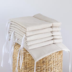 Peut inclure: Une pile de coussins carrés crème avec des liens en ruban blanc, posée sur un panier en osier. Les coussins semblent doux et matelassés, suggérant le confort et une palette de couleurs neutres pour divers styles de décoration.