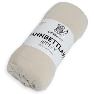 May include: A rolled-up, cream-coloured jersey knit fitted sheet. A white label wrapped around the sheet displays the text "CottonFast," "Pannbettlaken Jersey," and "100% Baumwolle" with a spool of thread graphic.
