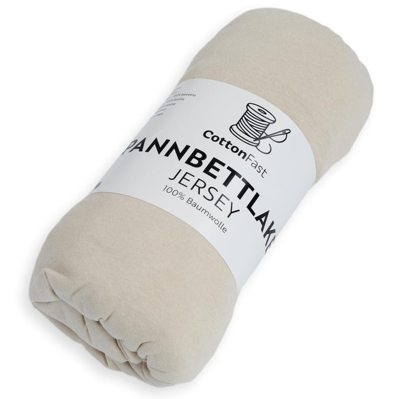 May include: A rolled-up, cream-coloured jersey knit fitted sheet. A white label wrapped around the sheet displays the text "CottonFast," "Pannbettlaken Jersey," and "100% Baumwolle" with a spool of thread graphic.