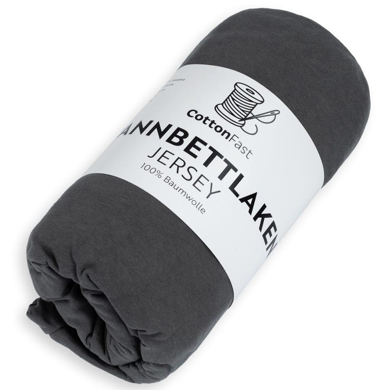 May include: Rolled-up dark grey jersey knit sheet. A white label displays the text "CottonFast" above a spool of thread graphic, and the text "Jannbettlaken Jersey 100% Baumwolle".