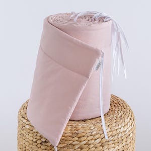Könnte beinhalten: Ein rosa Kinderbett-Stoßfänger mit weißen Bändern und dem Wort "LOOLAY" auf der Seite. Der Stoßfänger ist aufgerollt und liegt auf einem geflochtenen Korb.