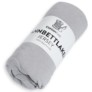 May include: A rolled-up, pale grey jersey knit sheet. A white label wraps around the sheet, displaying the text "CottonFast", a spool and needle graphic, and the words "WANNBETTLAKEN JERSEY 100% Baumwolle".