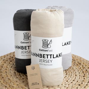 May include: Three rolled jersey bed sheets in grey, beige, and dark grey, each wrapped with a label featuring a spool of thread and the text "CottonFast ANNBETT LAKE JERSEY 100% Baumwolle". A small tag with text hangs in front.
