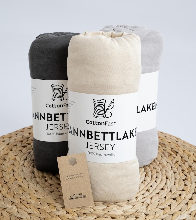 May include: Three rolled jersey bed sheets in grey, beige, and dark grey, each wrapped with a label featuring a spool of thread and the text "CottonFast ANNBETT LAKE JERSEY 100% Baumwolle". A small tag with text hangs in front.