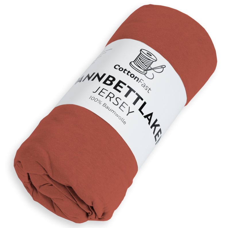 May include: Rolled terracotta-coloured jersey sheet. The sheet is wrapped with a white label that reads "CottonFast" and "Jannbettlake Jersey 100% Baumwolle". A spool of thread is on the label.