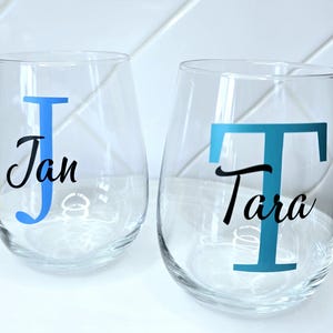 May include: Two clear glass wine glasses with personalised names. The glass on the left has a blue "J" and the name "Jan" in black. The glass on the right has a teal "T" and the name "Tara" in black.