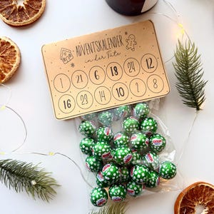 Può includere: Un'intestazione rettangolare in cartone per calendario dell'avvento con il testo "ADVENTSKALENDER in der Tüte" e cerchi numerati da 1 a 24, fissata a un sacchetto di plastica trasparente pieno di piccoli cioccolatini rotondi avvolti in carta verde a pois bianchi.