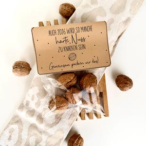 以下が含まれることがあります： 「AUCH 2026 WIRD SO MANCHE harte. Nuss ZU KNACKEN SEIN Gemeinsam packen wir das!」のテキストが書かれたクラフト紙のカードが木製のホルダーの上に置かれています。クルミは、クルミの入った透明な袋と模様の入った布の周りに散らばっています。
