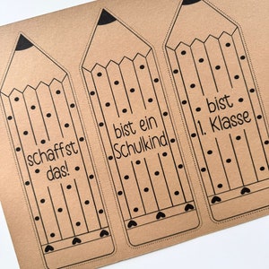 May include: Three brown paper pencil-shaped bookmarks with black outlines and polka dots. The bookmarks have the text "schafft das!" on the left, "bist ein Schulkind" in the middle, and "bist 1. Klasse" on the right.