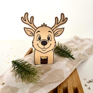 May include: A smiling reindeer-shaped gift box made of light brown cardboard. The reindeer has large antlers, a black nose, and a happy expression. The box is decorated with a small sprig of green pine and sits on a patterned beige and white runner.