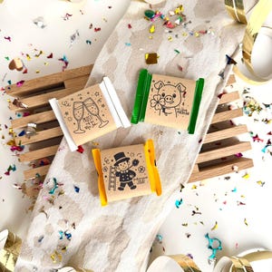 以下が含まれることがあります： お祝いのイラストとテキストが施された、3つの小さな包装されたギフトが、紙吹雪と金色のストリーマーが施された模様の布の上に配置されています。 ギフトには、シャンパングラス、豚、煙突掃除人の絵が描かれています。