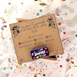 Op de afbeelding: Een bruine kaart met superheldenafbeeldingen en de Duitse tekst "ein paar Superkräfte FÜR DAS NEUE JAHR 2026". De kaart heeft cirkels met Duitse woorden en een mini-doosje Smarties. Confetti en lint omringen de kaart.