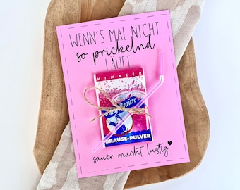 Può includere: Una carta rosa con testo tedesco e un piccolo pacchetto di "Himbeer Brause-Pulver" legato con spago. La carta, che recita "WENN'S MAL NICHT so prickelnd LAUFT" e "sauer macht lustig", è posta su un vassoio di legno con un panno fantasia.