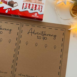 May include: A box of Kinder Chocolate with a child's face on the packaging, next to a brown paper with the words "Adventskranz to go" and other text. The image also includes star-shaped lights and a slice of dried orange.