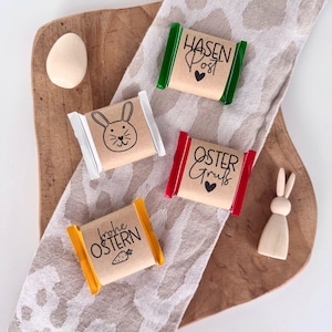 Könnte beinhalten: Drei kleine verpackte Schokoladentafeln mit Ostermotiven. Die Tafeln sind in braunes Papier mit bunten Rändern verpackt und zeigen Hasenillustrationen und Text in deutscher Sprache. Der Text auf den Tafeln lautet "Hasen Post", "Oster Gruss" und "Frohe Ostern".