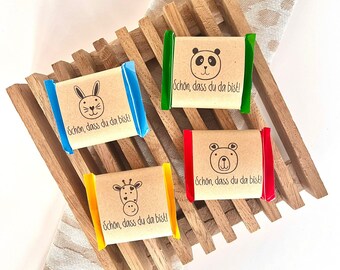 May include: Four small wrapped soaps with animal designs and the text "Schon, dass du da bist!" on each. The soaps are arranged on a wooden tray with a light brown fabric background.