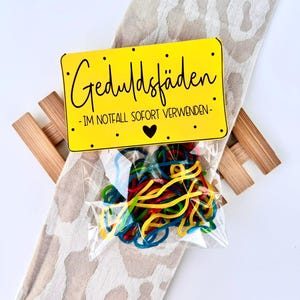 K&ouml;nnte beinhalten: Ein kleiner, durchsichtiger Plastikbeutel gef&uuml;llt mit bunten Gummib&auml;ndern, liegt auf einem beige-wei&szlig; gemusterten Stoff. Eine gelbe rechteckige Karte mit schwarzem Text "Geduldsf&auml;den - IM NOTFALL SOFORT VERWENDEN -" liegt oben auf dem Beutel.