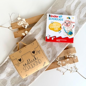 Può includere: Una scatola rettangolare di Kinder Schokolade con una pecora dei cartoni animati e il testo DU SCHA(F)FST DAS, accanto a una scatola regalo in cartone marrone chiaro con manico, stampata con il testo endlich SCHULKIND e icone scolastiche.