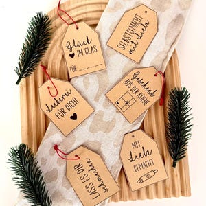Op de afbeelding: Een verzameling bruine papieren cadeaulabels met rode touwtjes, met handgeschreven tekst en kleine illustraties. De labels zijn gerangschikt op een houten dienblad met een patroonstof en takjes groen. De tekst bevat zinnen als "Glück im Glas" en "Mit Liebe Gemacht".