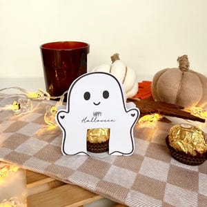Könnte beinhalten: Ein weißer Papier-Geister-Ausschnitt mit schwarzen Augen, einem kleinen Lächeln und dem aufgedruckten Text "Happy Halloween" auf der Vorderseite. Eine goldverpackte Schokoladenkugel befindet sich in einem Ausschnitt in der Mitte.