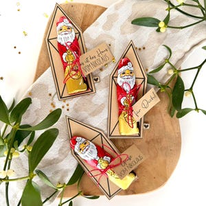 Pode incluir: Três figuras de chocolate do Pai Natal em embalagens vermelhas, cada uma com uma pequena etiqueta de presente. As etiquetas dizem "Grüße vom Nikolaus" e "für Dich!". Os chocolates estão numa tábua de madeira com vegetação.