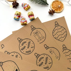 May include: A festive arrangement featuring brown paper sheets printed with black Christmas ornament designs. Accompanying the sheets are chocolate candies, dried orange slices, star anise, and a sprig of evergreen.