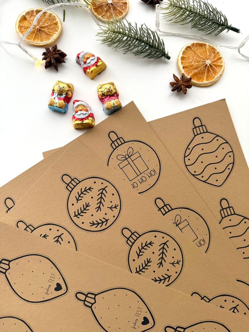 May include: A festive arrangement featuring brown paper sheets printed with black Christmas ornament designs. Accompanying the sheets are chocolate candies, dried orange slices, star anise, and a sprig of evergreen.