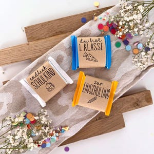 Puede incluir: Tres pequeños regalos envueltos con papel marrón y envoltorios coloridos. Los regalos tienen texto e ilustraciones, incluyendo una mochila, un cucurucho escolar y un lápiz. Los regalos están sobre una tela estampada con confeti y flores blancas.