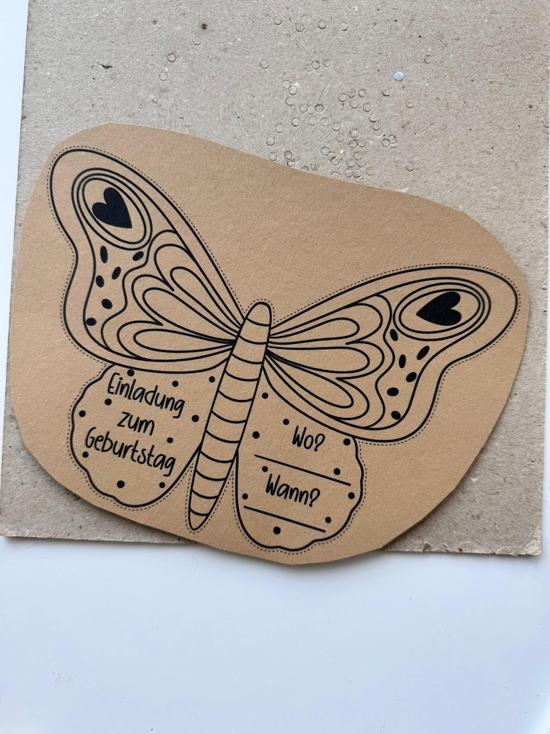 K&ouml;nnte beinhalten: Eine braune Papier-Einladung in Form eines Schmetterlings mit schwarzen Umrissen und Punkten. Der Text "Einladung zum Geburtstag" steht auf dem linken Fl&uuml;gel. Der rechte Fl&uuml;gel tr&auml;gt die Aufschrift "Wo?" und "Wann?"