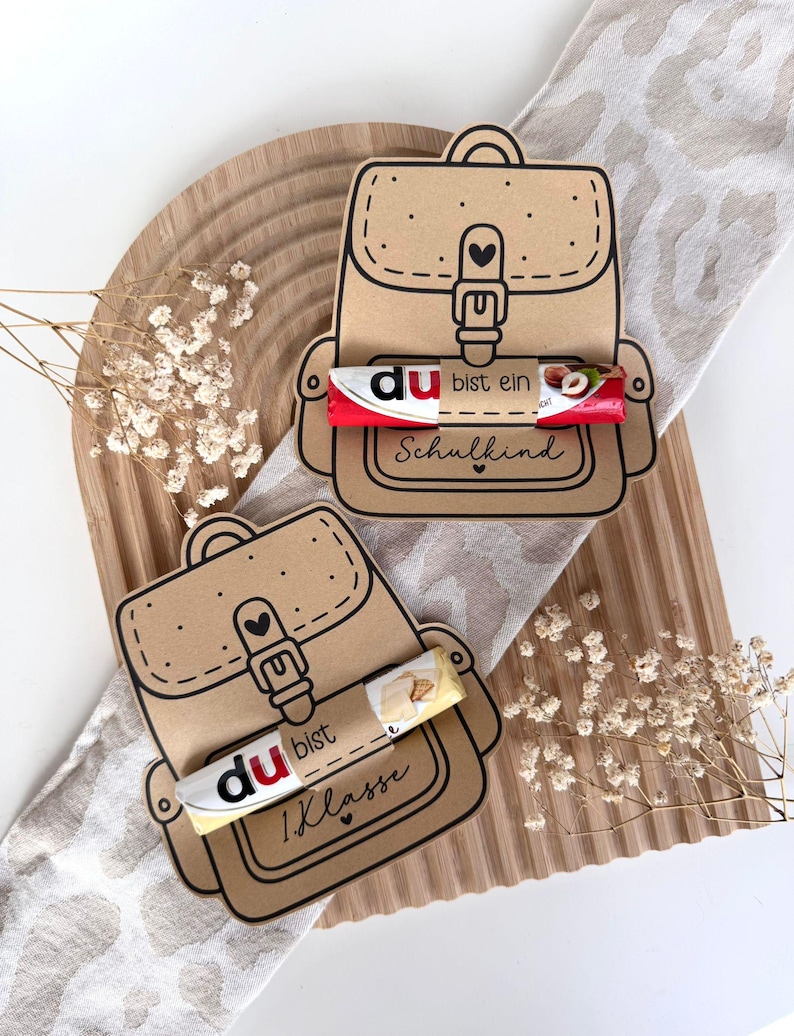 May include: Two brown paper backpack-shaped gift tags with black outlines, each holding a candy bar. One tag reads "du bist ein Schulkind" and the other reads "1. Klasse". The tags are on a wooden tray with dried flowers.