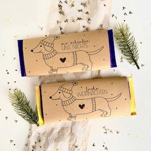 Op de afbeelding: Twee chocoladerepen verpakt in bruin papier, elk met een illustratie van een teckel met een hoed en trui. Op één staat "wir schenken UNS NICHTS" en op de andere "frohe WEIHNACHTEN". De folie is blauw en geel.