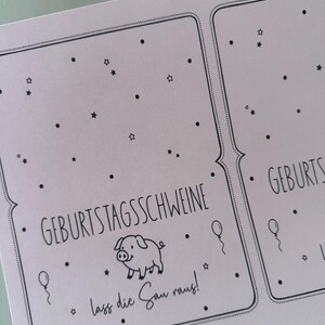 May include: A light pink card with black text and illustrations. The card features the words "GEBURTSTAGSSCHWEINE" and "lass die Sau raus!" along with a pig illustration, stars, balloons, and dots.