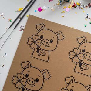 K&ouml;nnte beinhalten: Ein braunes Papier mit mehreren schwarz-wei&szlig;en Schweineaufklebern, die jeweils ein vierbl&auml;ttriges Kleeblatt und den Text "f&uuml;r DICH!" halten. Zwei Wunderkerzen und bunte Konfetti sind um das Papier verstreut.