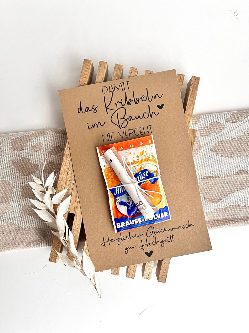 May include: A brown card with the text "DAMIT das Kribbeln im Bauch NIE VERGEHT" and a small packet of orange-flavored drink mix. The card also says "Herzlichen Gl&uuml;ckwunsch zur Hochzeit!" which translates to "Congratulations on your wedding!" in English.