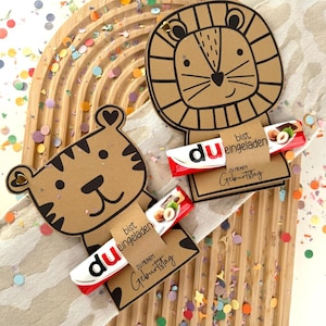 May include: Two brown cardboard animal-shaped gift tags, a tiger and a lion, each holding a candy bar. The tags have black outlines and the text "bist eingeladen" and "ZU MEINEM Geburtstag". Colorful confetti surrounds the tags.