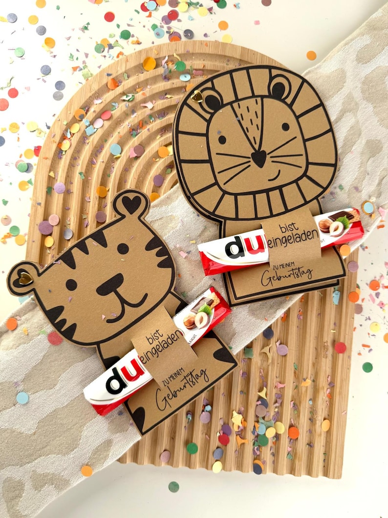 May include: Two brown cardboard animal-shaped gift tags, a tiger and a lion, each holding a candy bar. The tags have black outlines and the text "bist eingeladen" and "ZU MEINEM Geburtstag". Colorful confetti surrounds the tags.
