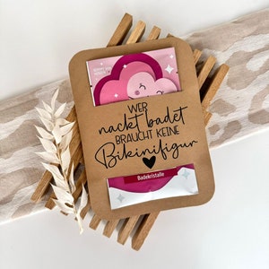 Peut inclure: Un sac cadeau en papier brun avec un motif de cœur rose et le texte "Wer nackt badet braucht keine Bikinifigur" et "Badekristalle".