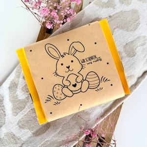 Op de afbeelding: Een ingepakt cadeau met een bruine papieren omslag met een cartoonkonijn met een hart, met de tekst "WIR SCHENKEN uns nichts". Het cadeau is verpakt in geel papier en is versierd met zwarte stippen. Roze bloemen en een houten standaard staan op de achtergrond.