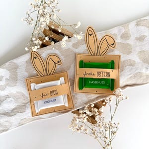 Può includere: Due scatole regalo a tema pasquale con orecchie da coniglio, ciascuna contenente una barretta di cioccolato. Una scatola dice "für DICH" e "JOGHURT", l'altra dice "Frohe Ostern" e "HASELNUSS". Le scatole sono marroni con dettagli verdi e bianchi.