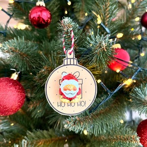 May include: A Christmas tree ornament featuring a Santa Claus design. The ornament is round with a light brown background and a Santa Claus illustration. The text "HO HO HO!" is printed below the Santa Claus image. The ornament is hanging on a Christmas tree.