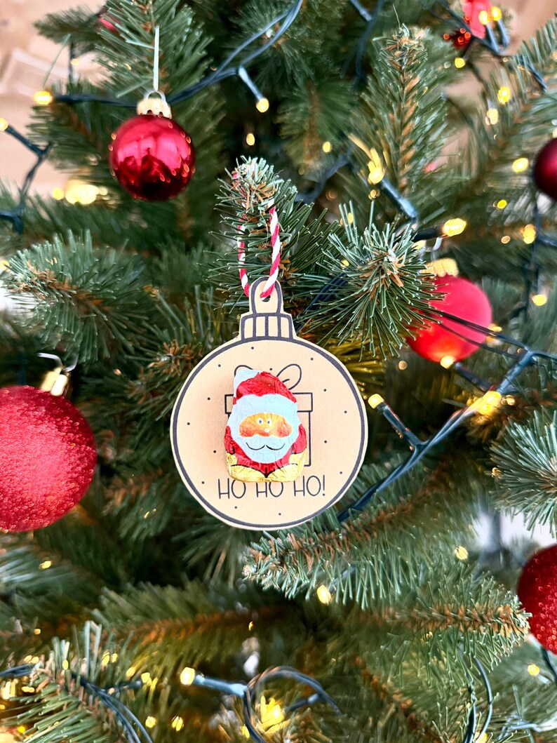 May include: A Christmas tree ornament featuring a Santa Claus design. The ornament is round with a light brown background and a Santa Claus illustration. The text "HO HO HO!" is printed below the Santa Claus image. The ornament is hanging on a Christmas tree.