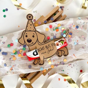 May include: A brown dachshund-shaped decoration with a party hat and the text "dackel gut ins neue Jahr!" is surrounded by confetti and gold ribbon. A red and white candy bar is attached to the dachshund. Wooden clothespins are in the background.