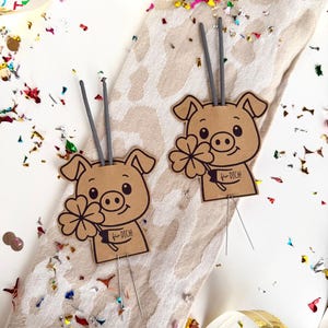 May include: Two brown pig-shaped party favors with a four-leaf clover and the text "für dich!" on a banner. Each pig has a gray stick attached. The favors are on a patterned beige cloth, surrounded by confetti and ribbons.