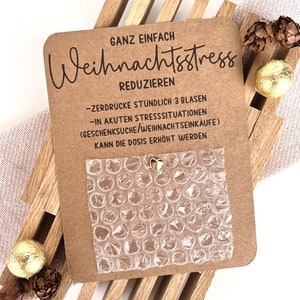 Könnte beinhalten: Eine braune Karte mit deutschem Text, darunter "Weihnachtsstress REDUZIEREN" und Anweisungen. Ein Blatt Luftpolsterfolie ist angebracht, mit einem kleinen goldenen Herzanhänger. Die Karte ist mit Holzlatten, Tannenzapfen und goldverpackten Pralinen verziert.