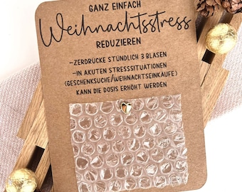 May include: A brown card with the German text "GANZ EINFACH Weihnachtsstress REDUZIEREN" and instructions. A sheet of bubble wrap is attached, with a small gold heart charm. The card is decorated with wooden slats, pine cones, and gold-wrapped chocolates.