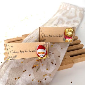 Op de afbeelding: Twee kleine kaarten met de tekst "Schön, dass du da bist!". Eén kaart heeft een chocoladefiguur van de kerstman, de andere een chocoladefiguur van een beer. De kaarten liggen op een beige servet met patronen, met houten accenten.
