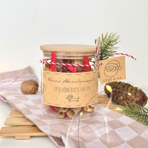 May include: A clear glass jar with a wooden lid containing rolled paper scrolls tied with red ribbon. A tan label reads "kleine Adventsauszeit 24 SCHÖNE BOTSCHAFTEN" with a dachshund icon. A tag features a Santa illustration and "GESCHENK für dich".